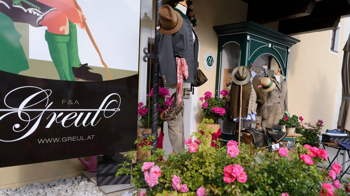 Ein storefront mit traditionellen Kleidungsstücken und Hüten, umgeben von bunten Blumen. Im Hintergrund ist ein großes Werbebanner sichtbar. | © Alexander Greul