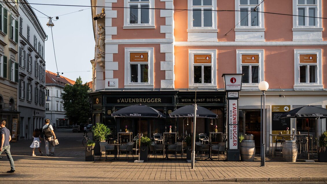 Ein gemütliches Restaurant mit Terrasse und Sonnenschirmen. Die Straße ist ruhig und von historischen Gebäuden umgeben. | © Auenbrugger
