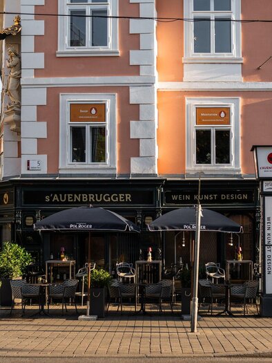 Ein gemütliches Restaurant mit Terrasse und Sonnenschirmen. Die Straße ist ruhig und von historischen Gebäuden umgeben. | © Auenbrugger