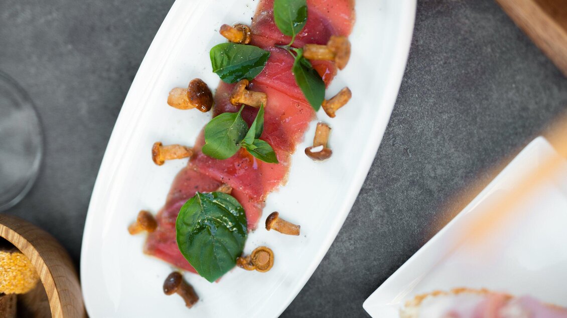 Ein elegantes Gericht mit dünn geschnittenem Fisch, garniert mit frischem Basilikum und Pilzen. Das Anrichten erfolgt auf einem langen weißen Teller. | © Auenbrugger