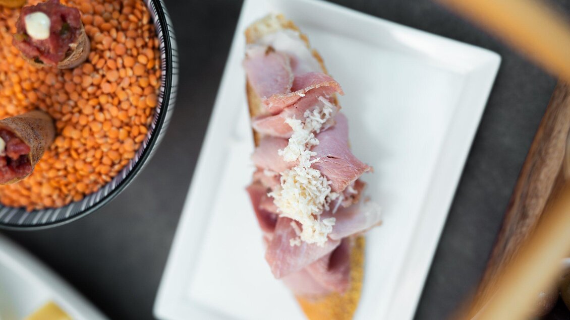 Ein Teller mit belegtem Brot, belegt mit Schinken und Käse. Daneben befindet sich eine Schüssel mit orangefarbener Linsen. | © Auenbrugger