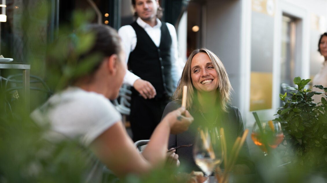 Eine fröhliche Unterhaltung zwischen zwei Frauen in einem Restaurant. Im Hintergrund ist ein Kellner zu sehen, der aufmerksam wirkt. | © Auenbrugger