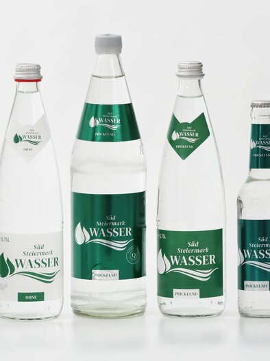 Five bottles of water in different sizes are standing against a white background. Next to the bottles is a glass with clear water. | © Aufhauser