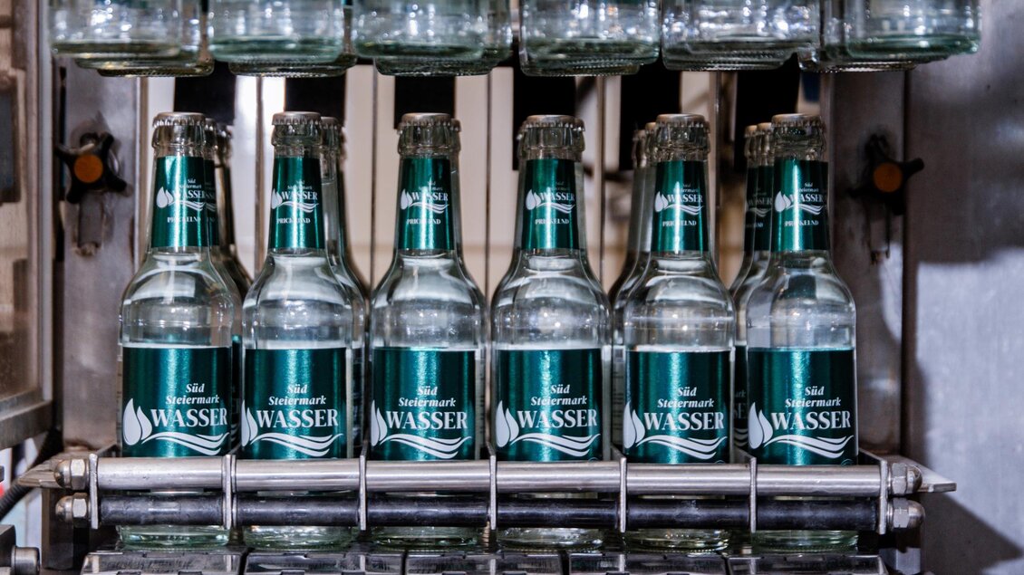 Eine Reihe von Flaschen mit Wasser steht auf einem Förderband. Die Flaschen sind klar und tragen ein Etikett mit der Aufschrift „WASSER“. | © RM SW GmbH | webquartier.at
