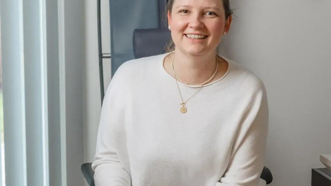 Eine lächelnde Frau sitzt in einem modernen Raum und hält eine Taschenlampe in der Hand. Sie trägt einen hellen Pullover und wirkt freundlich und einladend.