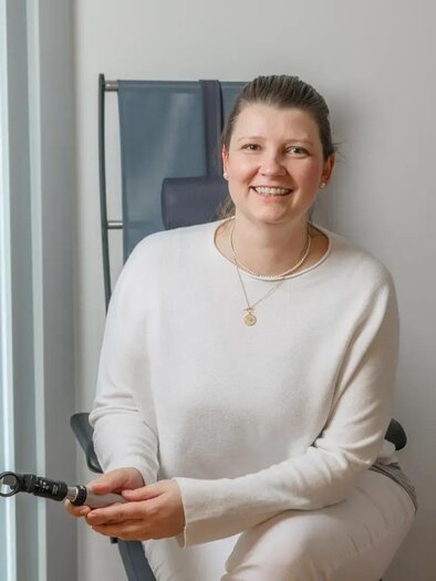 A smiling woman is sitting in a modern room and holding a flashlight in her hand. She is wearing a light sweater and appears friendly and inviting.