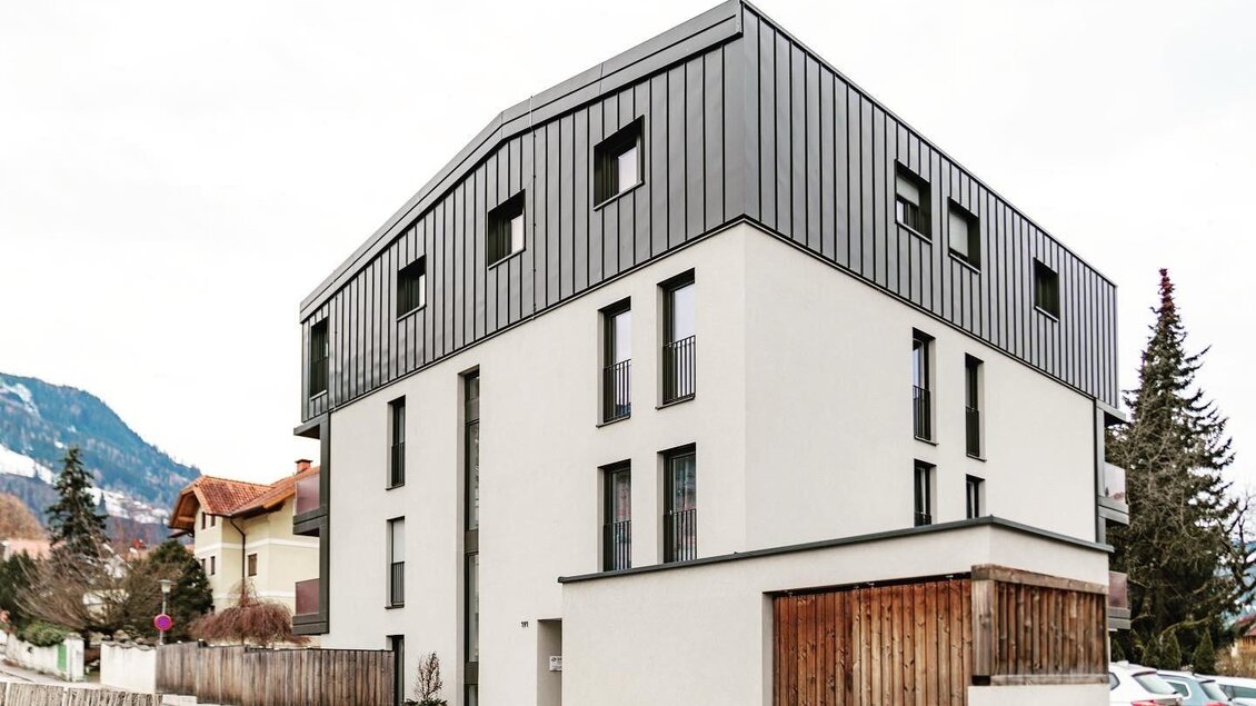 Ein modernes Gebäude mit einer Kombination aus grauer und weißer Fassade. Im Hintergrund sind Berge und Bäume zu sehen.