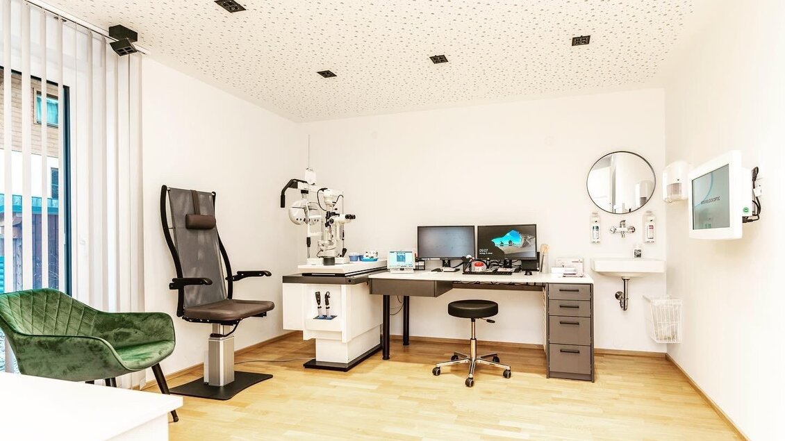 Ein modernes Untersuchungszimmer mit einem komfortablen Stuhl und medizinischer Ausstattung. Der Raum ist hell und gut organisiert, mit einem Schreibtisch und einem Monitor.