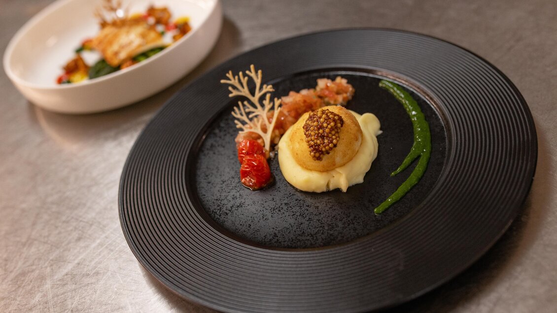 Ein elegantes Gericht auf einem schwarzen Teller, bestehend aus püriertem Gemüse, Kaviar und einer Garnitur aus Kräutern. Im Hintergrund ist ein weiteres Gericht in einer weißen Schüssel sichtbar.