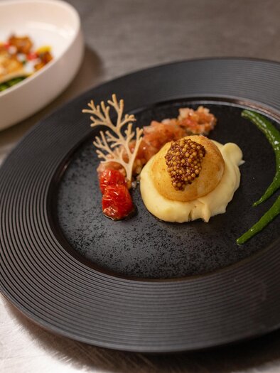 An elegant dish on a black plate, consisting of pureed vegetables, caviar, and a garnish of herbs. In the background, another dish is visible in a white bowl.
