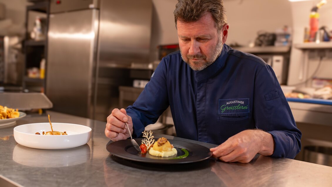 Ein Koch, der sorgfältig ein Gericht auf einem schwarzen Teller anrichtet. Im Hintergrund sind Küchengeräte und ein weiterer Teller mit Essen sichtbar. | © 5komma5Sinne