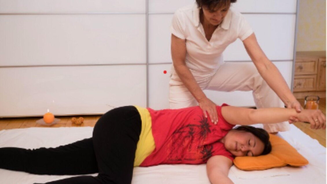 Eine Massage, bei der eine Frau einer anderen Frau den Rücken und die Arme behandelt. Entspannende Atmosphäre mit einem orangefarbenen Kissen und sanftem Licht. | © Johanna Erhart