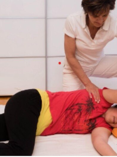 Eine Massage, bei der eine Frau einer anderen Frau den Rücken und die Arme behandelt. Entspannende Atmosphäre mit einem orangefarbenen Kissen und sanftem Licht. | © Johanna Erhart