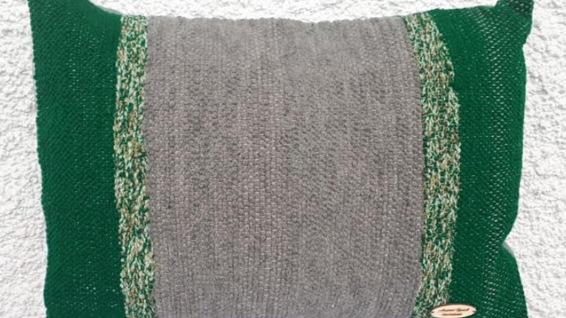 Ein dekorives Kissen in grauer und grüner Farbe mit einem strukturierten Muster. Es hat ein einfaches, modernes Design und einen kleinen Markenanhänger. | © Ausseer Teppich_Melanie Schlömmicher