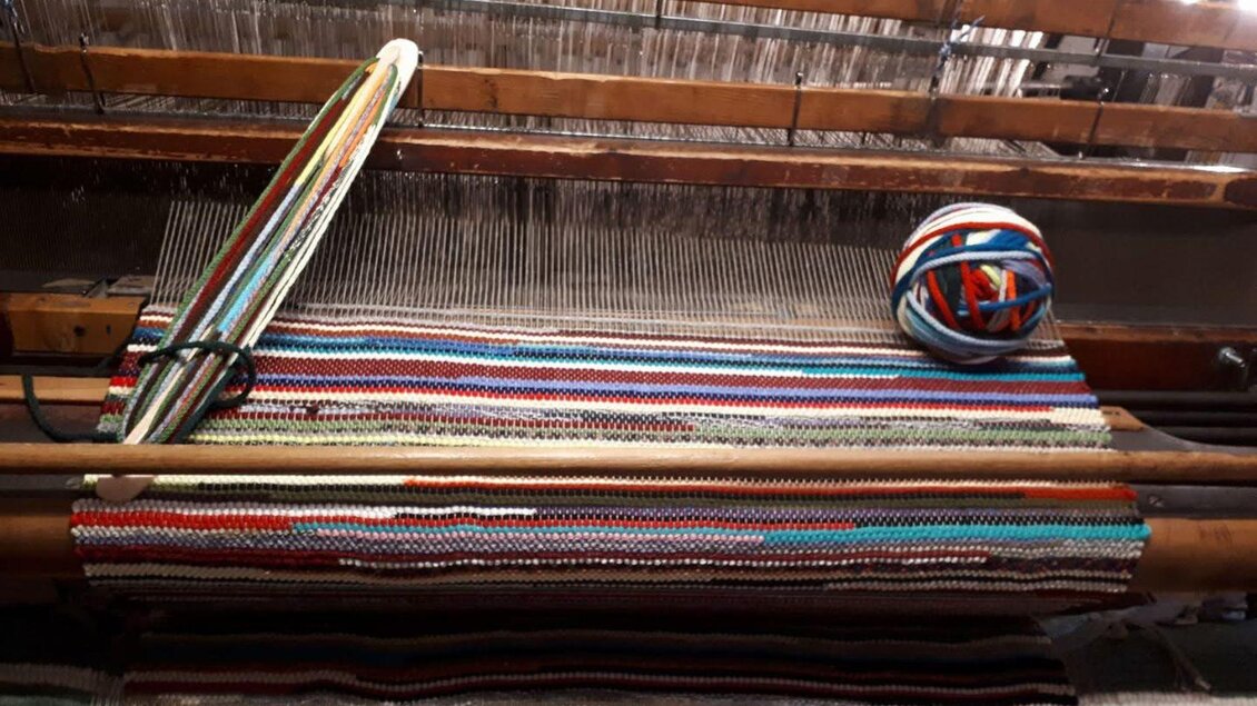 Ein Webstuhl mit bunten Fäden und einem farbigen Garnknäuel. Die Streifen im Gewebe zeigen verschiedene Farben und Muster. | © Ausseer Teppich