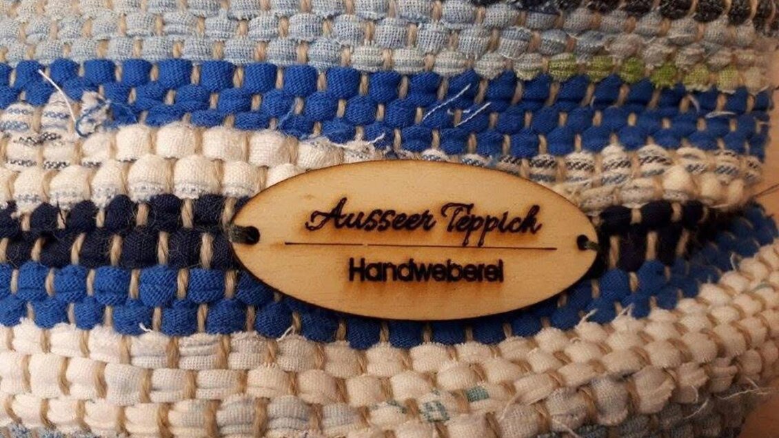Ein handgewebter Teppich mit blauen und weißen Streifen. Das Etikett zeigt den Namen "Ausseer Teppich" und die Bezeichnung "Handweberei". | © Ausseer Teppich