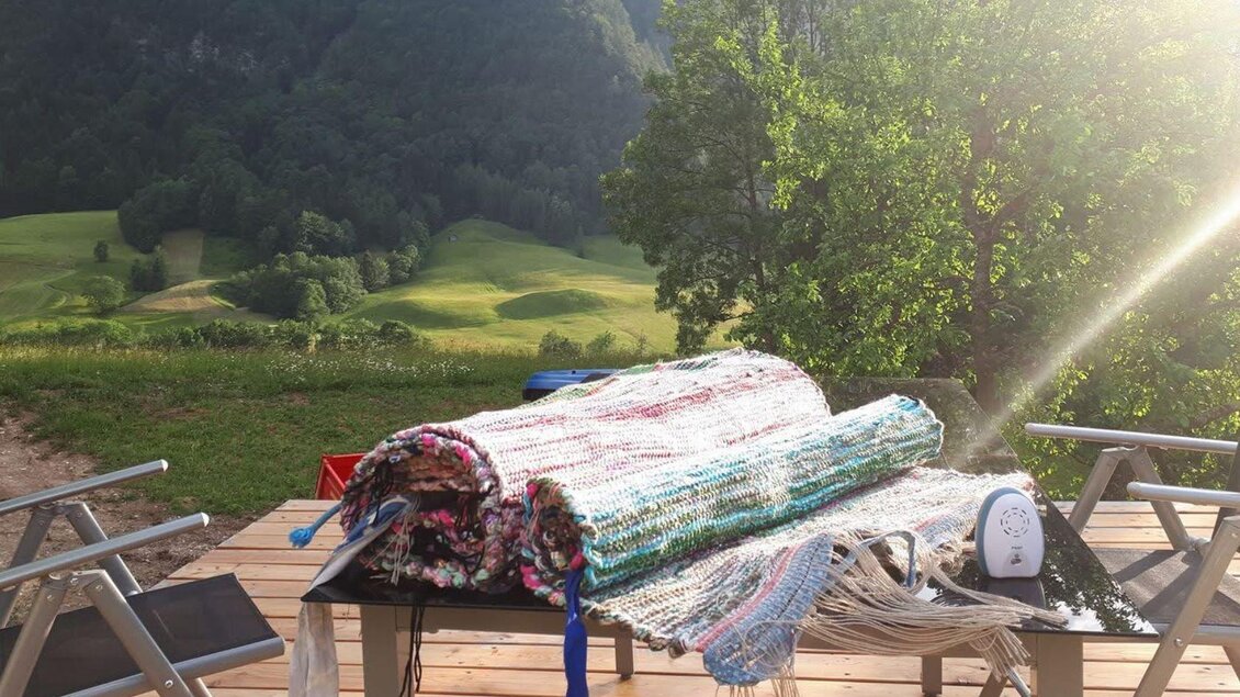 Ein Tisch auf einer Terrasse mit bunten Teppichen und einem Bügeleisen. Im Hintergrund sind sanfte Hügel und Bäume zu sehen. | © Ausseer Teppich