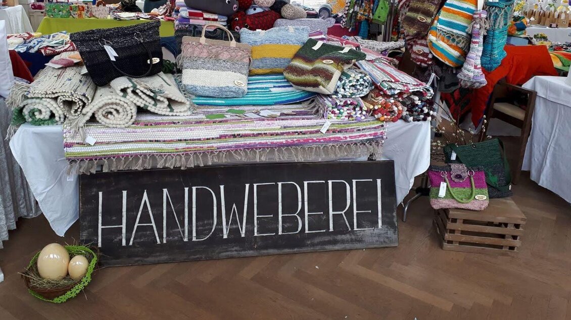 Ein Tisch voller handgefertigter Textilien, darunter Decken und Schals. Ein Schild mit der Aufschrift "HANDWEBEREI" steht prominent im Vordergrund. | © Ausseer Teppich