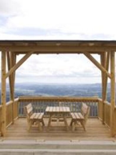 Eine Holzveranda mit Tisch und Bänken bietet einen Blick auf die Landschaft. Die Umgebung ist grün und hügelig unter einem bewölkten Himmel. | © Aussichtswarte Buchenberg