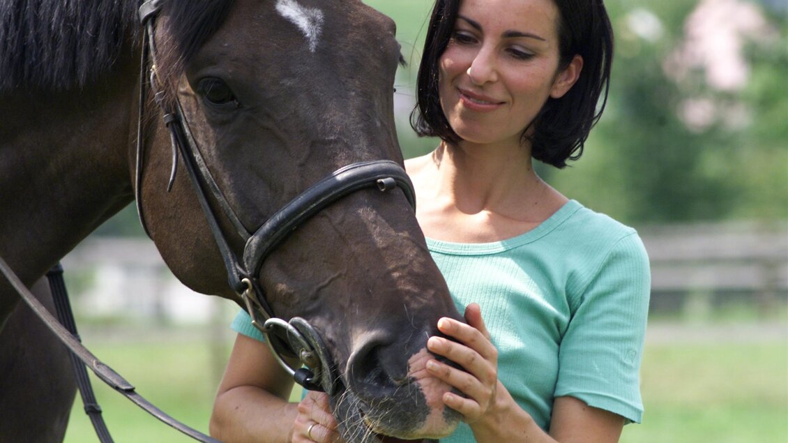 Eine Frau steht neben einem Pferd und streichelt es liebevoll. Im Hintergrund sind sanfte Hügel und ein Gebäude zu sehen. | © Kurkommission Bad Blumau