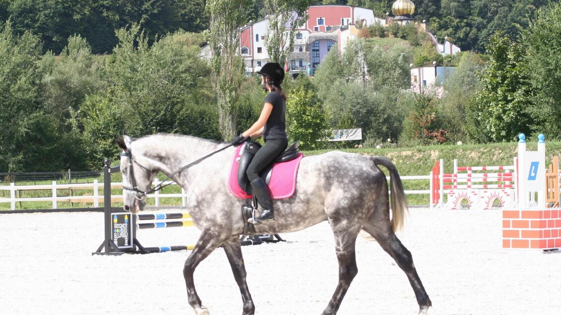 Eine Reiterin sitzt auf einem grauen Pferd und reitet in einem Dressurviereck. Im Hintergrund sind Bäume und einige Gebäude sichtbar. | © Kurkommission Bad Blumau