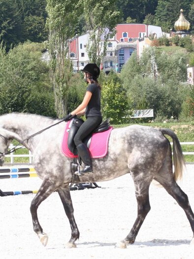 Eine Reiterin sitzt auf einem grauen Pferd und reitet in einem Dressurviereck. Im Hintergrund sind Bäume und einige Gebäude sichtbar. | © Kurkommission Bad Blumau