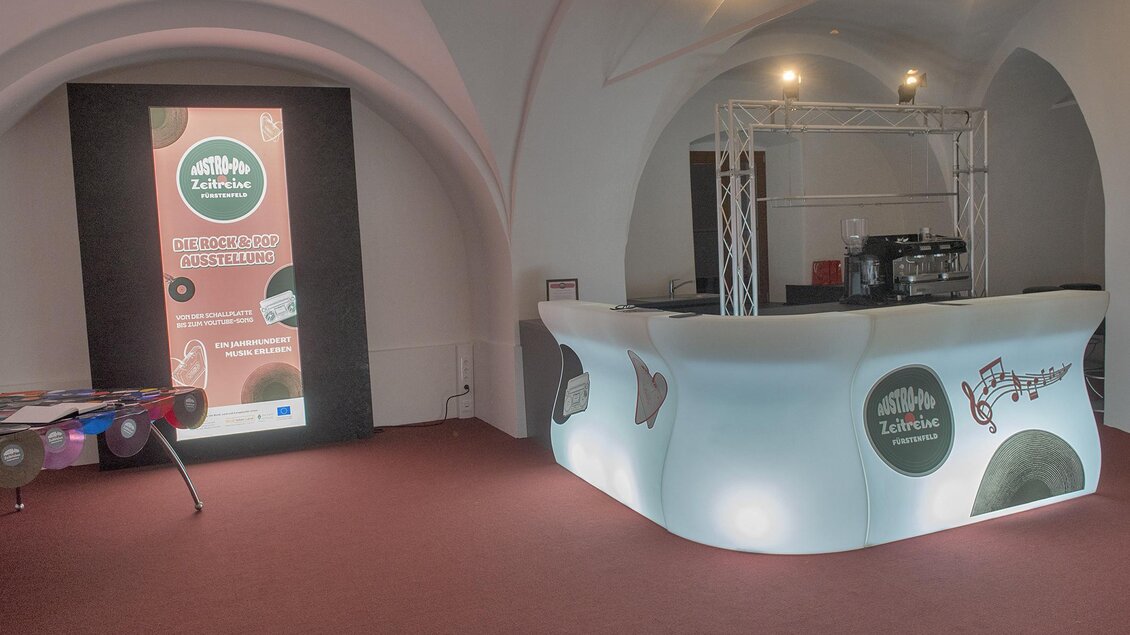 Ein modern gestalteter Empfangsbereich mit einem beleuchteten Tresen. Im Hintergrund befindet sich ein großer Bildschirm mit Werbung. | © Austro-Pop-Zeitreise