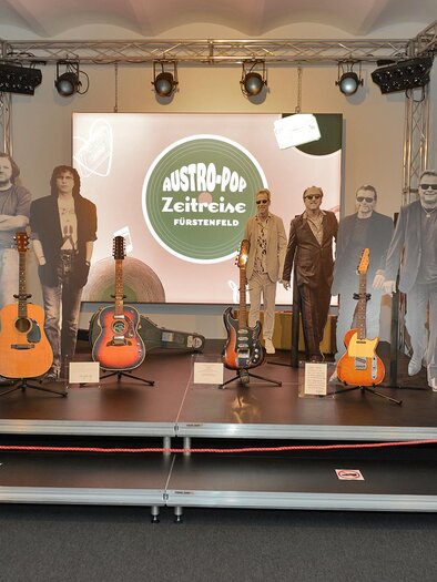 Eine Ausstellung mit Gitarren und lebensgroßen Figuren aus Papier. Im Hintergrund ist ein großes Bildschirm mit dem Schriftzug "Austro-Pop Zeitreise". | © Austro-Pop-Zeitreise