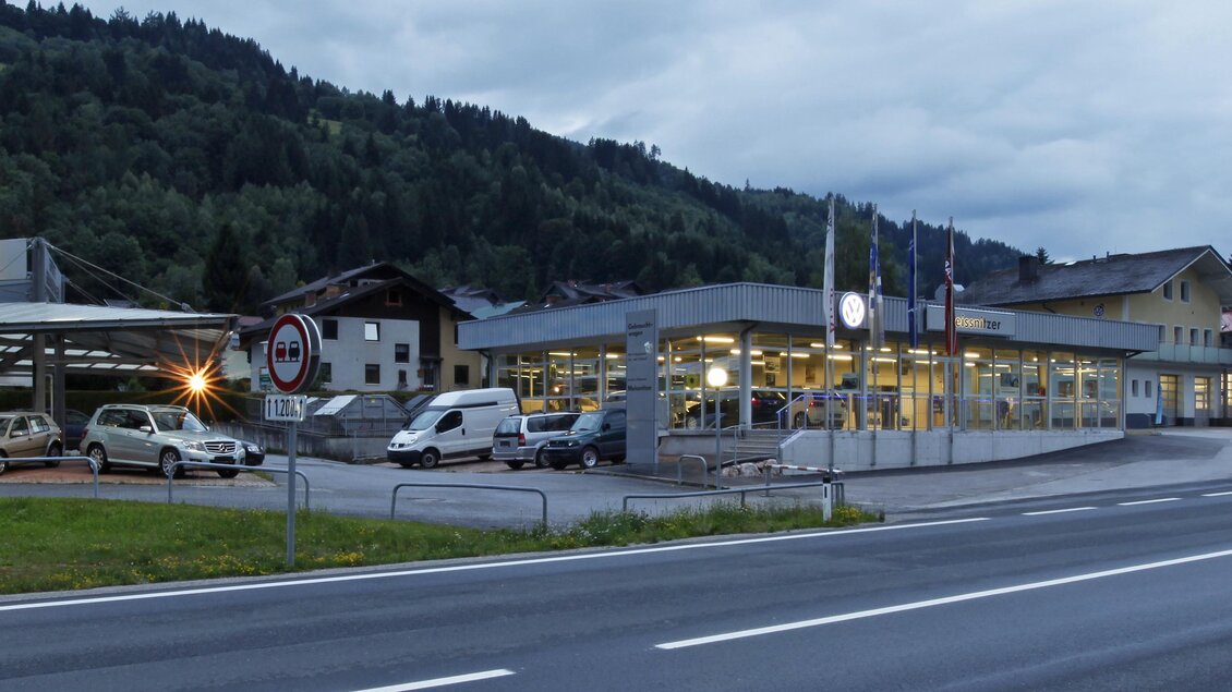 Ein Autohaus mit mehreren Fahrzeugen auf dem Parkplatz steht an einer Straße. Im Hintergrund sind sanfte Hügel und bewaldete Flächen zu sehen. | © Autohaus Meissnitzer