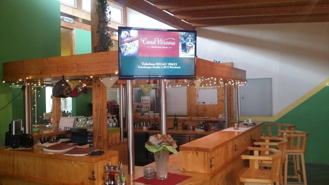 Ein gemütlicher Innenraum mit einer Holzbar und Stühlen. Ein Bildschirm zeigt Werbung für ein Restaurant an der Wand. | © keine Kenntnis