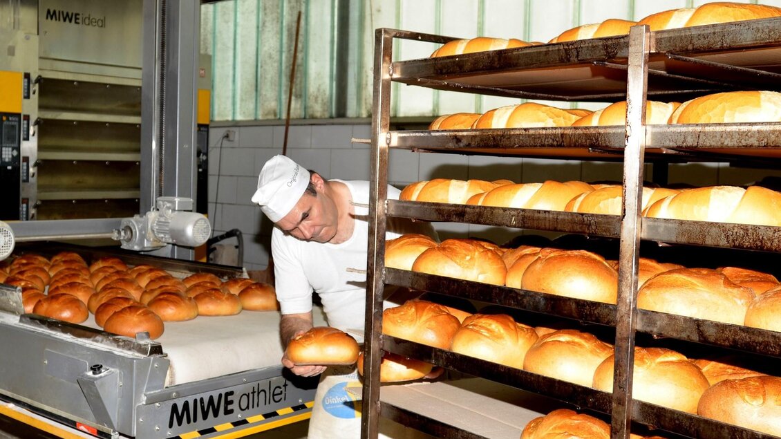 Ein Bäcker arbeitet in einer Bäckerei und überprüft frisch gebackenes Brot. Im Hintergrund sind Regale mit weiteren Broten zu sehen. | © Bäckerei & Konditorei Wolf