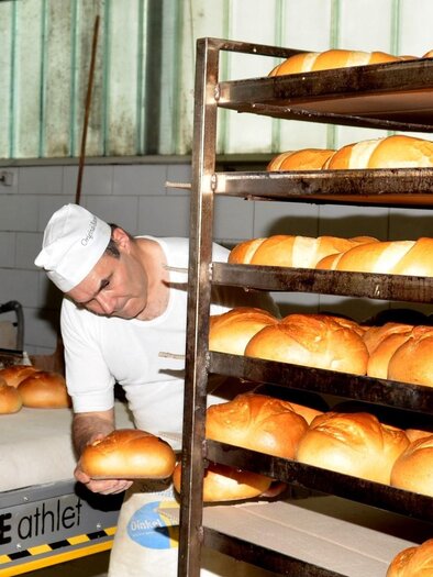Ein Bäcker arbeitet in einer Bäckerei und überprüft frisch gebackenes Brot. Im Hintergrund sind Regale mit weiteren Broten zu sehen. | © Bäckerei & Konditorei Wolf