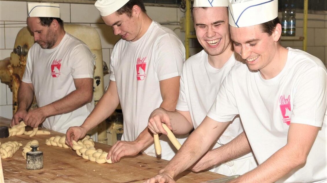 Vier junge Bäcker arbeiten gemeinsam an einem Tisch und formen Teig. Alle tragen weiße Shirts und Kochmützen und lachen dabei. | © Bäckerei & Konditorei Wolf