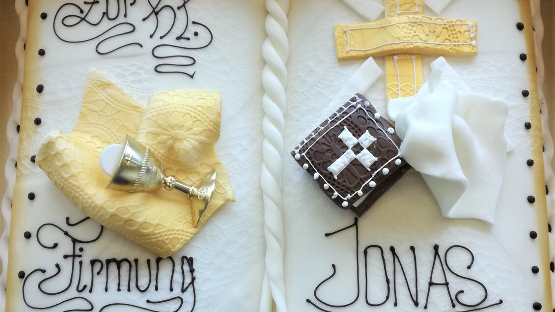 Eine festliche Torte in Buchform mit dem Namen "Jonas". Auf der Torte sind religiöse Symbole, darunter ein Kreuz, eine goldene Krone und ein Kelch, schön gestaltet. | © Bäckerei & Konditorei Wolf