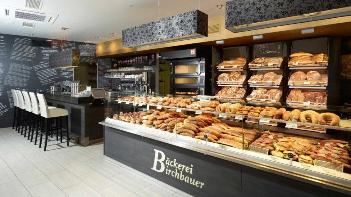 Eine moderne Bäckerei mit einer großen Auswahl an frischem Brot und Gebäcken. Im Hintergrund steht eine Theke mit Barhockern. | © Bäckerei Birchbauer