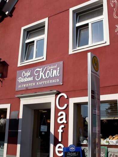 Ein charmantes Café mit roter Außenwand und großen Fenstern. Über der Tür steht "Café Blütenblatt Könii," ein einladender Ort für Kaffeeliebhaber. | © C. Stein