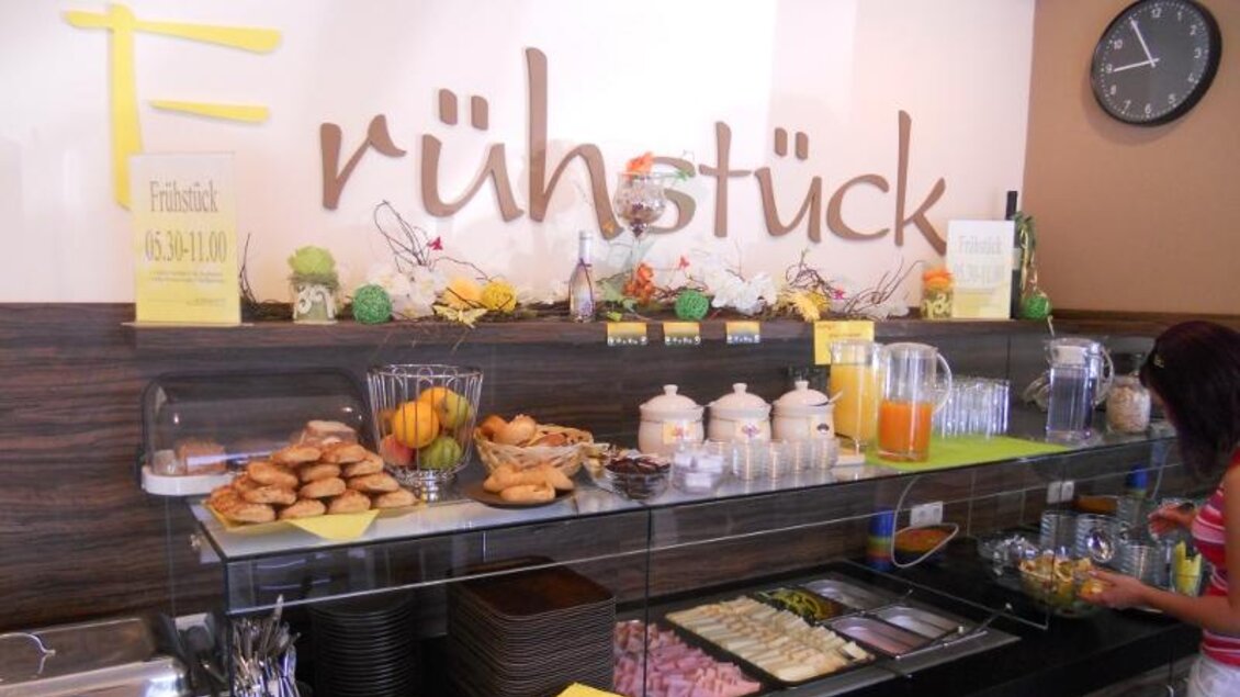 Ein Frühstücksbuffet mit verschiedenen Speisen, darunter Brötchen, Aufschnitt und Obst. Im Hintergrund steht eine Uhr und eine dekorierte Wand mit der Aufschrift „Frühstück“. | © Bäckerei Konditorei Café Hütter