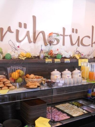 Ein Frühstücksbuffet mit verschiedenen Speisen, darunter Brötchen, Aufschnitt und Obst. Im Hintergrund steht eine Uhr und eine dekorierte Wand mit der Aufschrift „Frühstück“. | © Bäckerei Konditorei Café Hütter