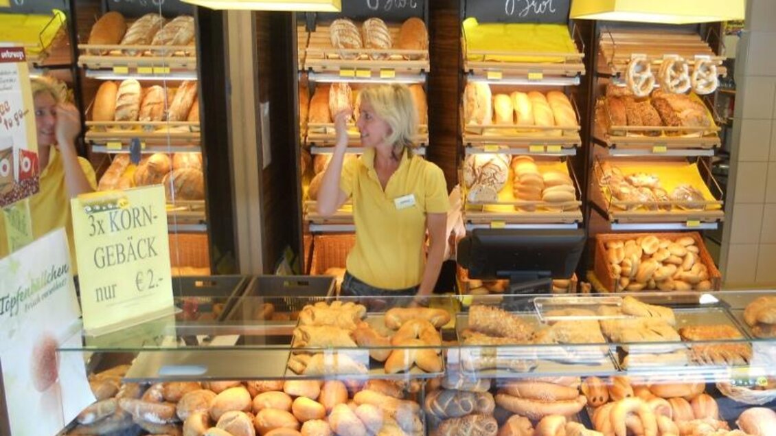 Eine Bäckerei mit einer großen Auswahl an Brötchen und Gebäck. Eine Verkäuferin in einem gelben Shirt steht hinter der Theke und bedient Kunden. | © Bäckerei Konditorei Café Hütter