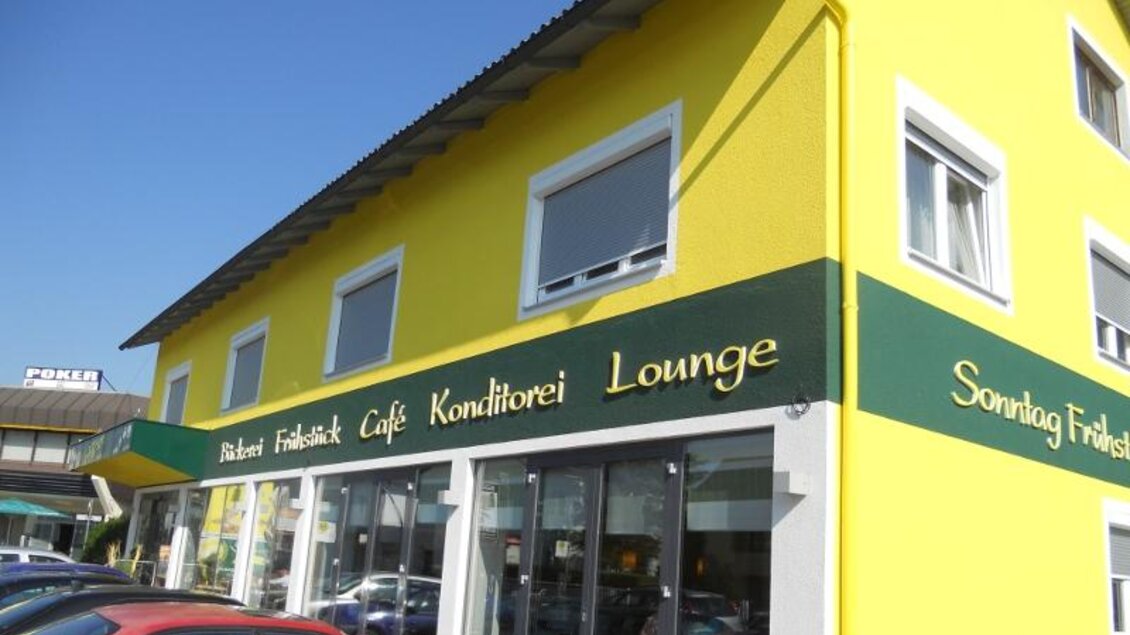 Ein buntes Café mit gelber Fassade und einer einladenden Lounge. Die Fenster sind groß und bieten einen Blick auf die Straße. | © Bäckerei Konditorei Café Hütter