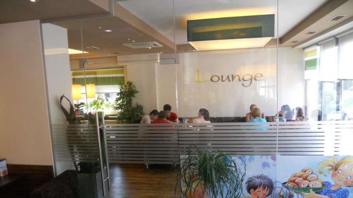 Eine gemütliche Lounge mit mehreren Personen, die am Tisch sitzen. Der Raum ist hell und modern gestaltet, mit Pflanzen und einer Wandverkleidung. | © Bäckerei Konditorei Café Hütter