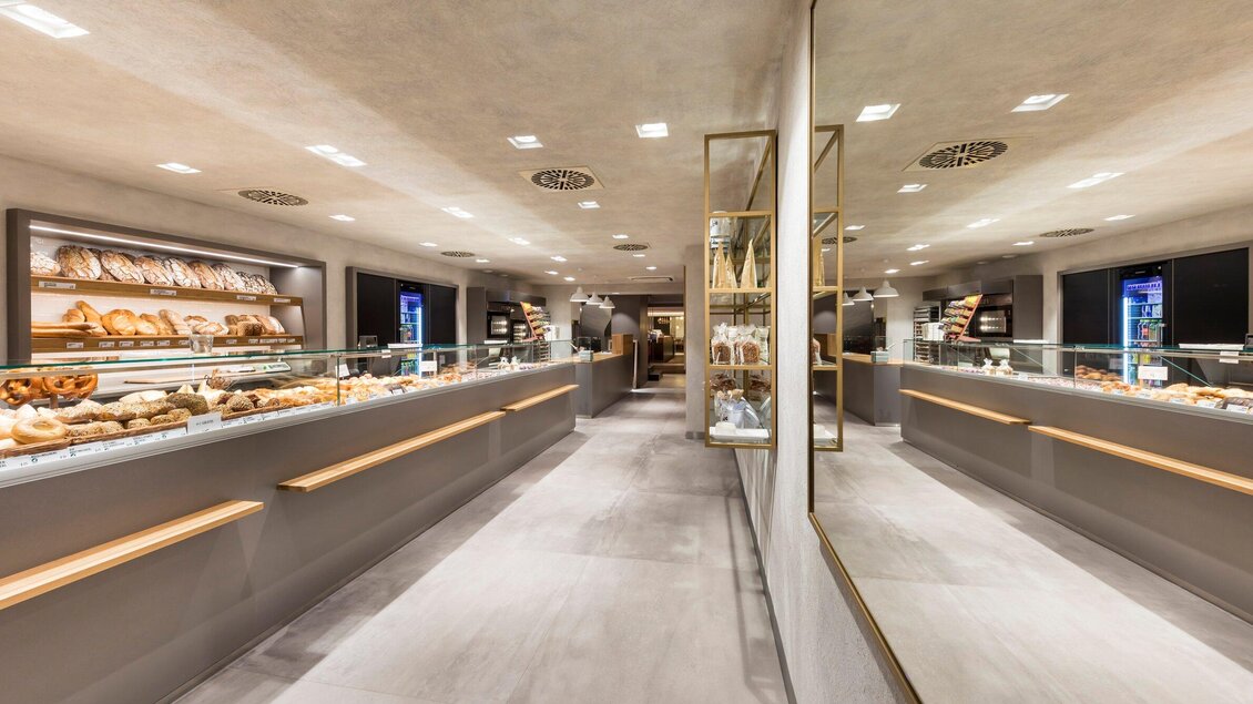 Ein modernes Café oder Bäckerei mit hellen Wänden und eleganten Regalen. Überall sind frisch gebackene Brötchen und Croissants zu sehen. | © Sorger - Werner Krug