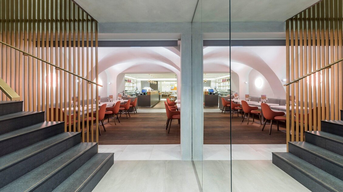 Ein modernes Restaurant mit eleganter Innenausstattung und sanften Farben. Die Treppe führt zu einem einladenden Speisebereich mit roten Stühlen. | © Sorger - Werner Krug