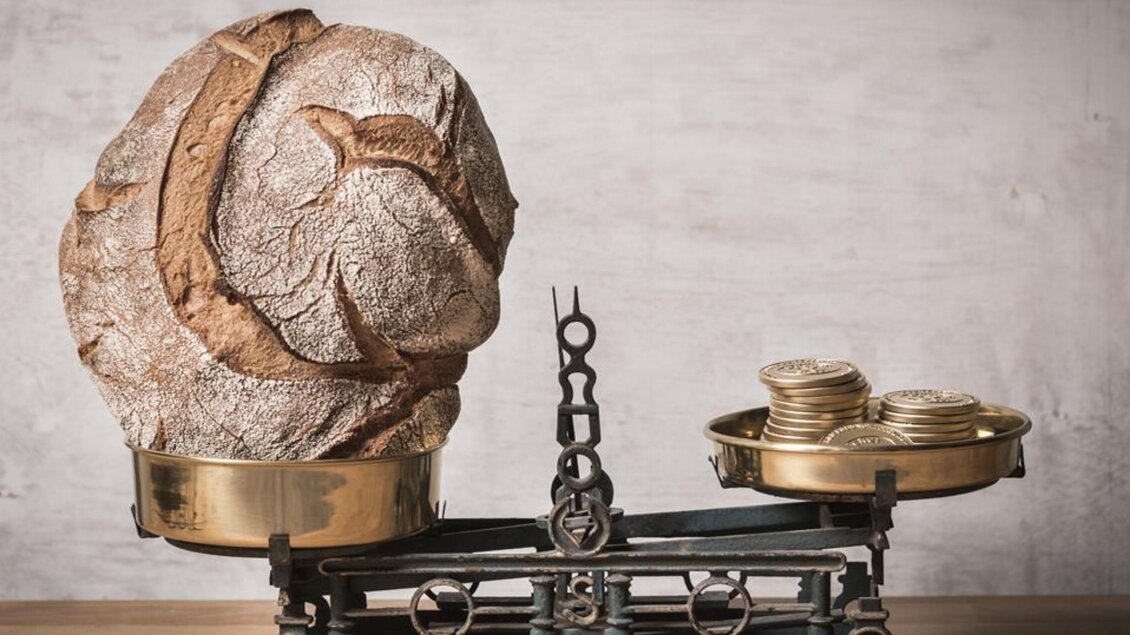 Ein großes Laib Brot auf einer alten Balance, die auf der linken Seite überwiegt. Auf der rechten Seite der Waage befinden sich einige Münzen. | © Werner Krug