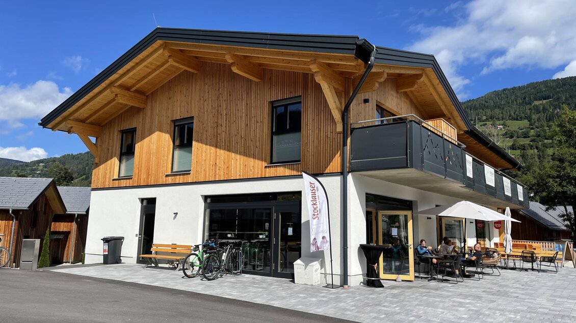 Ein modernes, zweigeschossiges Gebäude mit Holzverkleidung und einem gemütlichen Außenbereich. Der Himmel ist blau und es sind einige Tische draußen auf der Terrasse. | © Tourismusverband Murau