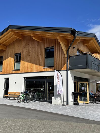 Ein modernes, zweigeschossiges Gebäude mit Holzverkleidung und einem gemütlichen Außenbereich. Der Himmel ist blau und es sind einige Tische draußen auf der Terrasse. | © Tourismusverband Murau