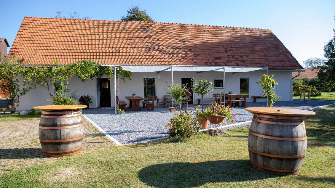 Ein gemütliches Haus mit rotem Dach und überdachter Veranda. Im Vordergrund stehen zwei Fass-Tische und grünes Gras. | © Bäcksteffl Bauernspezereien