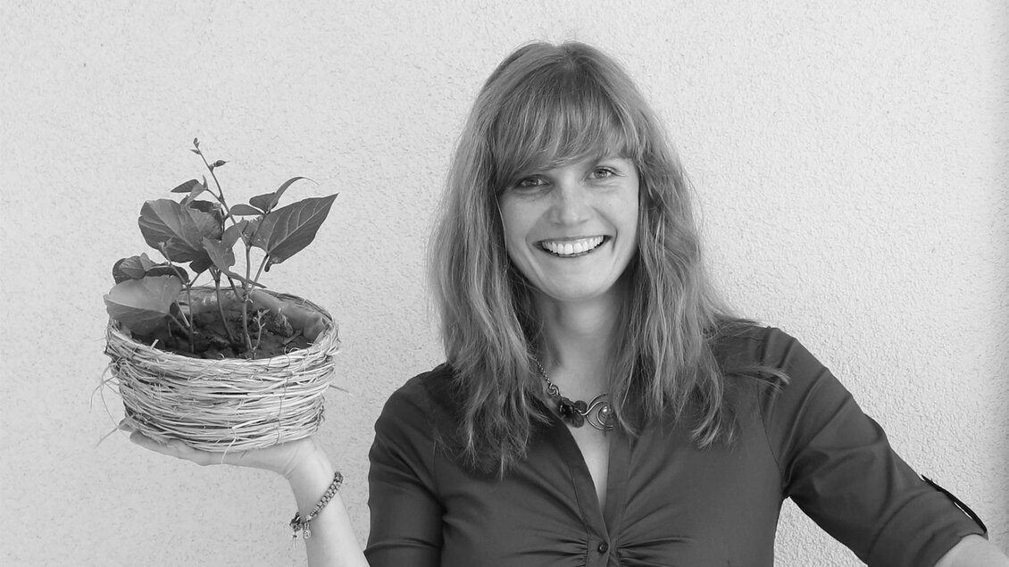 Eine lächelnde Frau hält einen Pflanzenkübel in der Hand. Der Kübel ist aus geflochtenem Material und enthält eine kleine Pflanze. | © Bäcksteffl