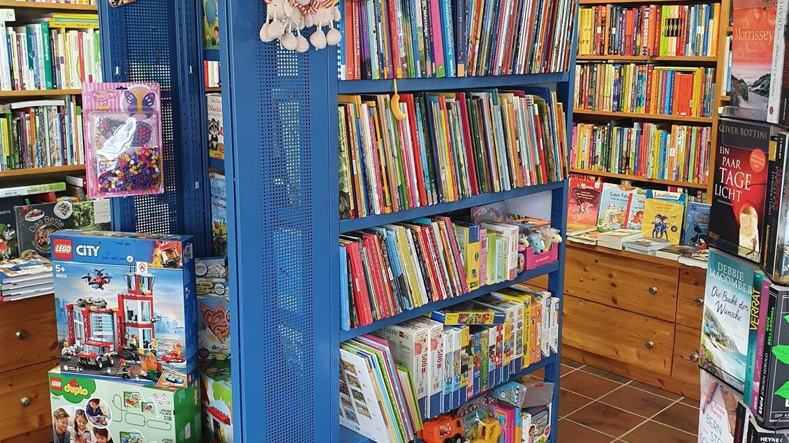 Ein Bücherregal in einem Buchladen, gefüllt mit bunten Kinderbüchern. Plüschtiere hängen oben am Regal, und rundherum sind weitere Bücher und Spielzeuge zu sehen. | © Büchereck Barbara Freidinger