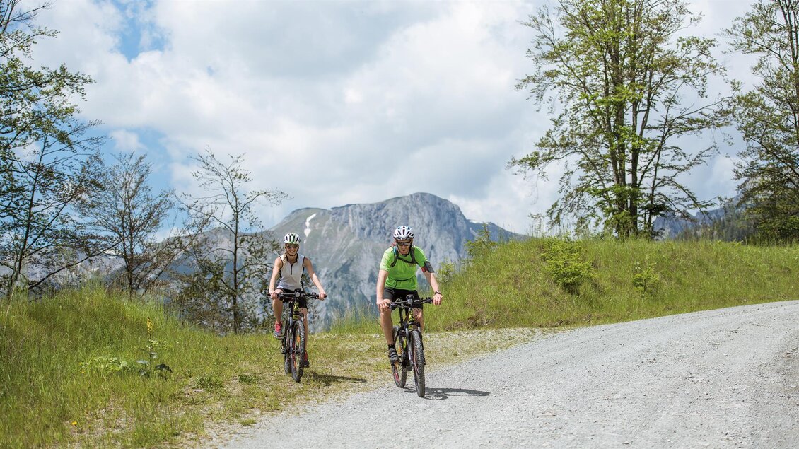 Zwei Radfahrerinnen fahren einen Schotterweg entlang. Im Hintergrund sind Berge und ein blauer Himmel mit Wolken zu sehen.